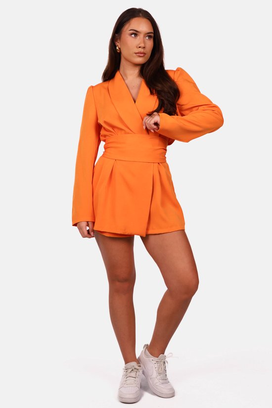 Model in oranje blazerjumpsuit met witte sneakers, gefotografeerd tegen een witte achtergrond.