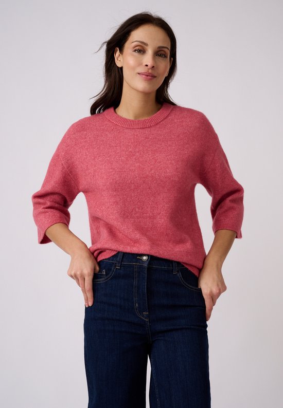 Een model draagt een roze gebreide trui met wijde driekwartmouwen, gecombineerd met een donkerblauwe spijkerbroek.