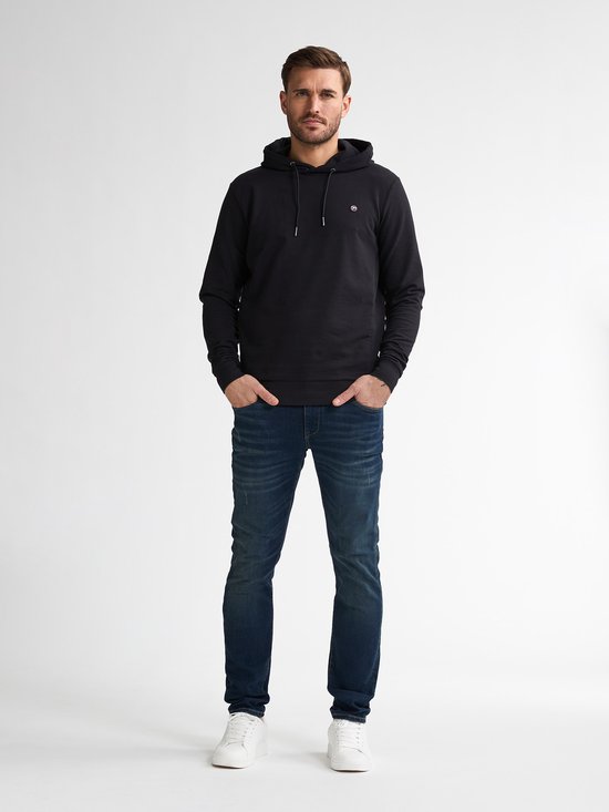 Man in donkerblauwe slim fit jeans en zwart hoodie, met witte sneakers.