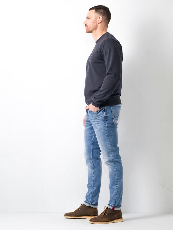 Man in donkere trui en blauwe regular tapered fit jeans met stretchdenim. Bruine schoenen.