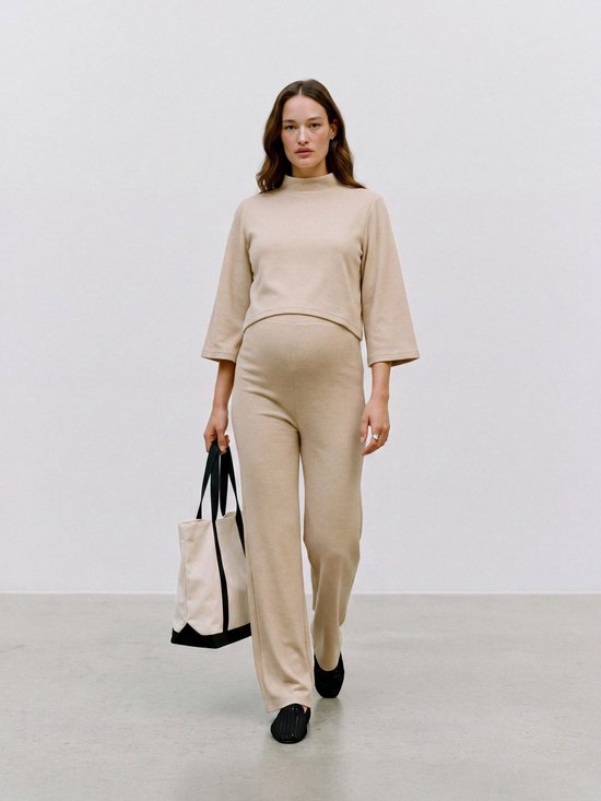 Een zwangere vrouw in een beige lounge set, bestaande uit een cropped top en broek met wijde pijpen. Ze draagt een tas en zwarte schoenen.