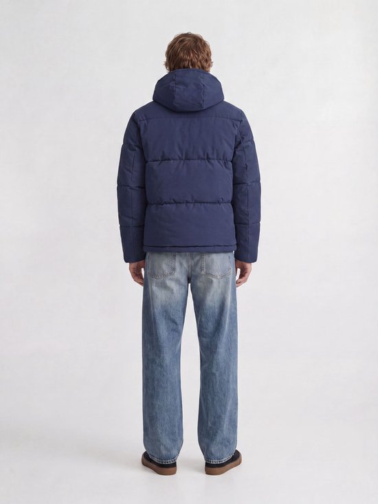 Man met de rug naar voren, draagt een blauwe pufferjas met capuchon en wijde jeans.