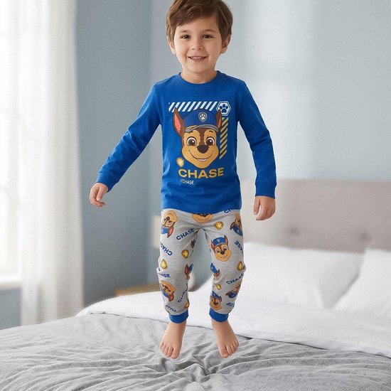 Jongetje springt op een bed in een blauwe pyjama met Chase van Paw Patrol op de borst en patroon van Chase op de broek.