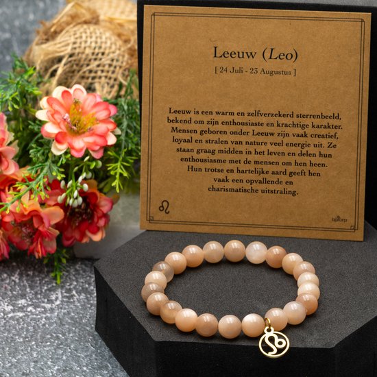Armband met perzikkleurige zonnesteen kralen en gouden Leo-bedel, naast een kaart en bloemen.