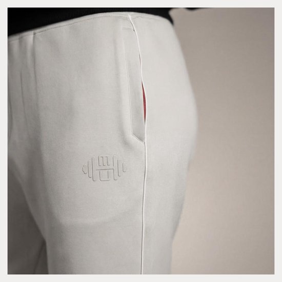 Close-up van de zijkant van de beige broek met witte bies, rode zakdetail en reliëflogo op de heup.