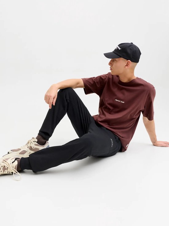 Model draagt zwarte joggingbroek, donkerrood T-shirt met 'Originals Studio' opdruk en beige sneakers.