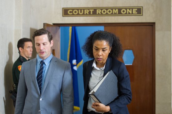 Een man en vrouw lopen uit de rechtszaal. De vrouw draagt een map. Boven hen staat 'COURT ROOM ONE'.