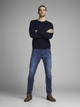 Man met gekruiste armen in een donkerblauwe trui en blauwe jeans met een lichte wassing.