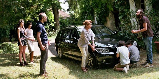 Een groep mensen staat buiten bij een zwarte auto in de zon. Een man met baard praat met een vrouw.