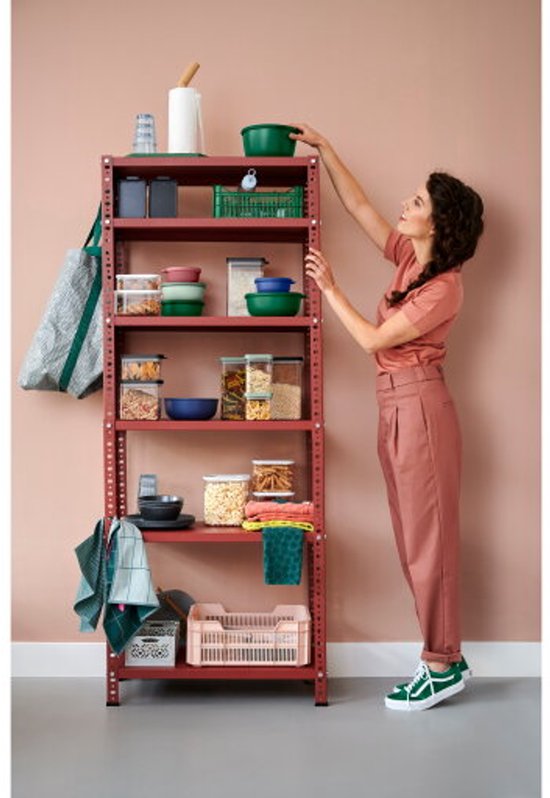 Une femme en tenue rose place un bol vert sur une étagère de rangement rouge avec diverses boîtes de conservation Mepal et d'autres articles de cuisine.