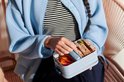 Personne en veste bleue prend un sandwich d'une boîte à lunch bleu clair avec des poivrons rouges.