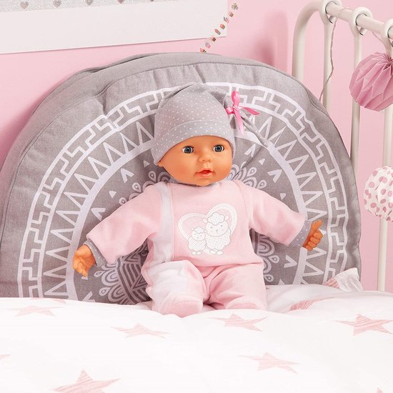 Babypop in roze slaappakje met grijze stippenmuts, zittend op een bedje.
