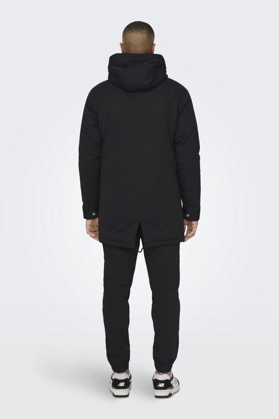 Achterkant van zwarte parka met capuchon, gedragen door een man in zwarte broek en sneakers.