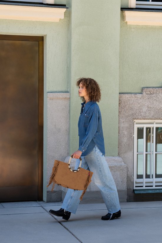 Model loopt met een bruine suède tas met franjes en gouden handvatten. Ze draagt een spijkerblouse en jeans.
