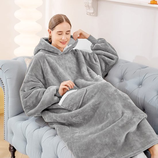 Vrouw ontspant op een bank in een comfortabele, grijze, oversized hoodie deken met witte fleece voering.