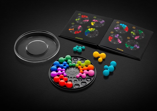 IQ Circle spel op zwart oppervlak. Gekleurde, ronde stukken liggen in en naast het spelbord. Een open boek met opdrachten ligt ernaast.