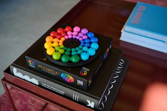 Gekleurde, ronde stukken van het IQ Circle spel liggen op een zwart spelbord, dat op een stapel boeken ligt. De stukken vormen een regenboogpatroon.