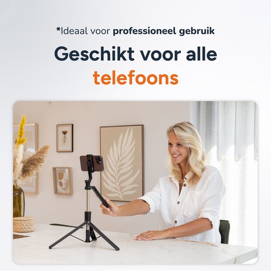Vrouw met blond haar, witte blouse en spijkerbroek, staand bij een bureau met een selfiestick.