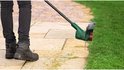 Persoon in zwarte broek en laarzen maait gras met groene grastrimmer langs een stenen pad.