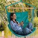 Vrouw in blauwe trui en jeans drinkt uit mok in turquoise hangstoel, met palmbomen op achtergrond.