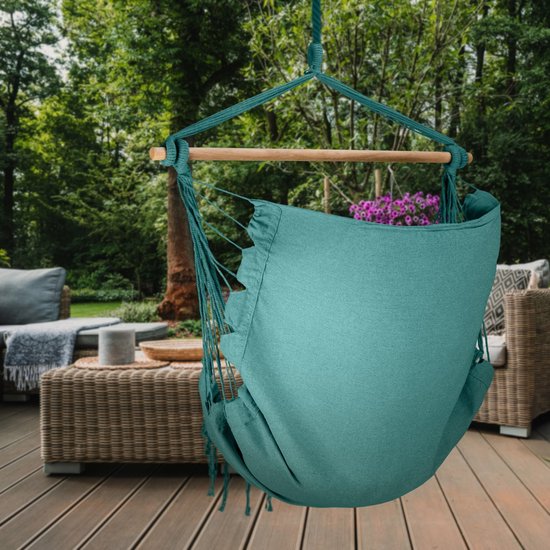 Turquoise hangstoel met franjes en houten spreidstok, opgehangen op een houten terras met tuinmeubilair.