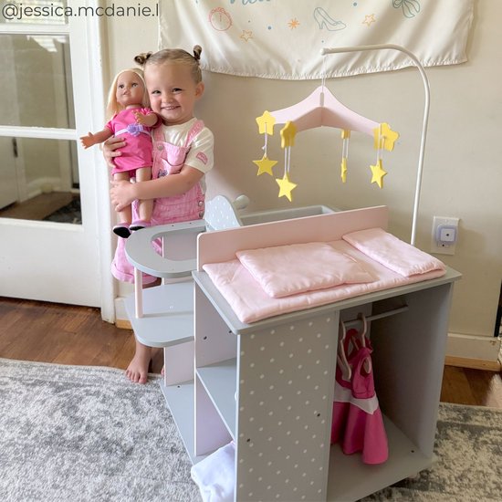 Meisje met staartjes in een roze tuinbroek houdt een pop vast. Rechts staat een grijze commode met roze details en een mobiel met sterren.