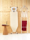 Crèmekleurige strijkplank rechtop met ophangbeugel, naast een kledingrek met kleding.
