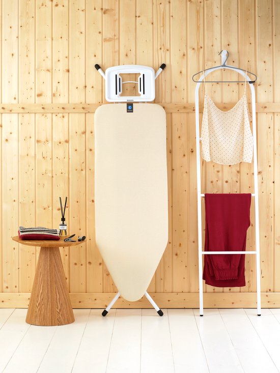 Crèmekleurige strijkplank rechtop met ophangbeugel, naast een kledingrek met kleding.