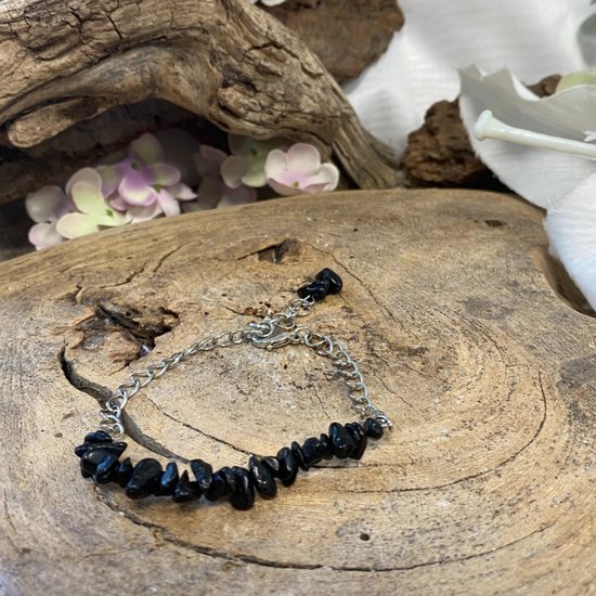 Zilverkleurig schakelarmbandje met ruwe zwarte steentjes, tentoongesteld op een stuk hout met achtergrondbloemen.