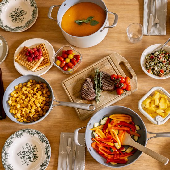 Houten tafel met diverse pannen en borden gevuld met maaltijden, waaronder biefstuk, salade en soep.