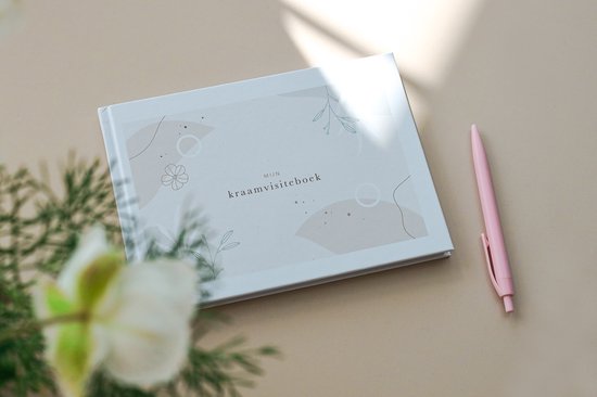 Een gesloten kraamvisiteboek met een licht beige omslag, versierd met abstracte vormen en botanische illustraties. Een roze pen ligt ernaast, met een bloem op de voorgrond.