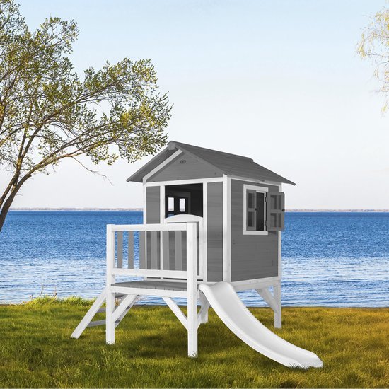 Houten speelhuisje met grijze wanden, verdieping en witte glijbaan, bij een meer.