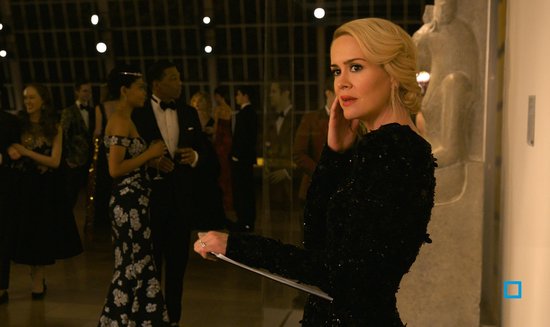 Sarah Paulson in een zwarte, glinsterende avondjurk, met gasten op een gala-evenement op de achtergrond.