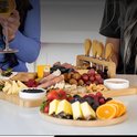 Personen genieten van een borrelplank gevuld met fruit, kaas en worst, terwijl een vrouw een glas witte wijn vasthoudt.