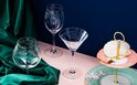 Un verre à champagne, un verre à vin et un verre à martini avec des gravures complexes, sur fond rose et bleu.