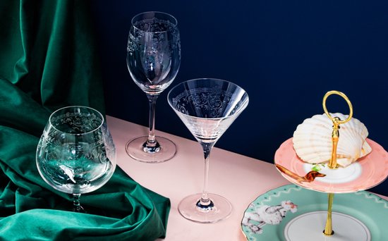 Un verre à champagne, un verre à vin et un verre à martini avec des gravures complexes, sur fond rose et bleu.