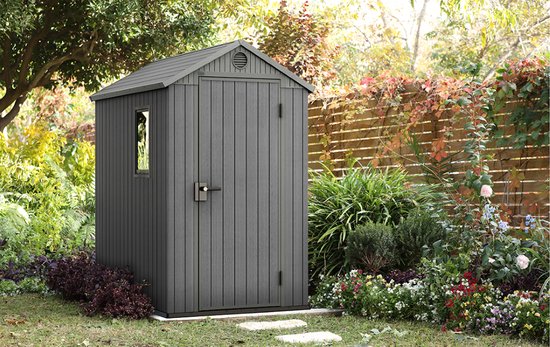 Grijze berging met gesloten deuren, raam en ventilatie in een tuin met bloemen.