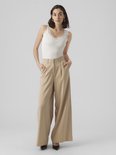 Model staat met de hand in de zak van een beige wijde broek en draagt een wit kanten hemdje.