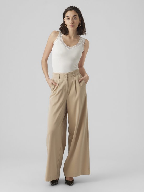 Model staat met de hand in de zak van een beige wijde broek en draagt een wit kanten hemdje.