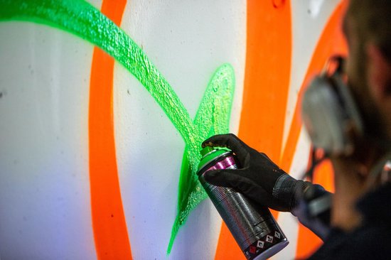 Hand met spuitbus met felgroene verf, spuitend op oranje cirkels op een witte muur.