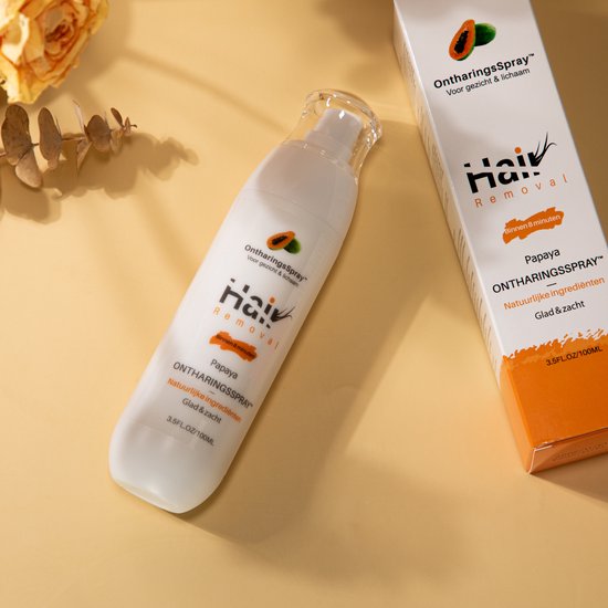Witte fles en doosje ontharingsspray met papaja-logo, op een houten schijf met gedroogde bladeren.
