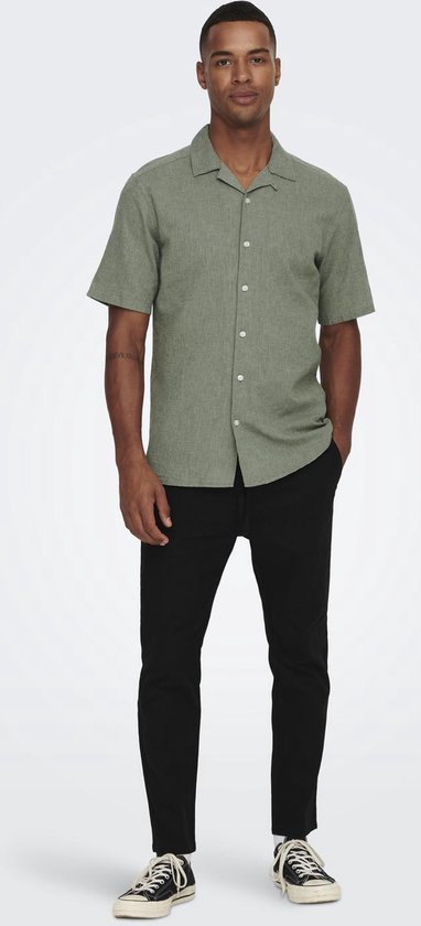 Man draagt groen, kortemouwige hemd met linnen textuur, wit T-shirt en zwarte broek.
