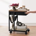 Zijtafel met houten blad, een zwarte stoffen mand, een metalen frame met wieltjes en een netwerkplank. Gevuld met boeken en een koptelefoon.