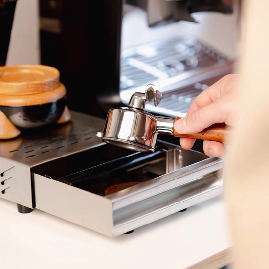 Een hand houdt een zilverkleurige portafilter met houten handvat boven een uitklaplade van geborsteld staal.