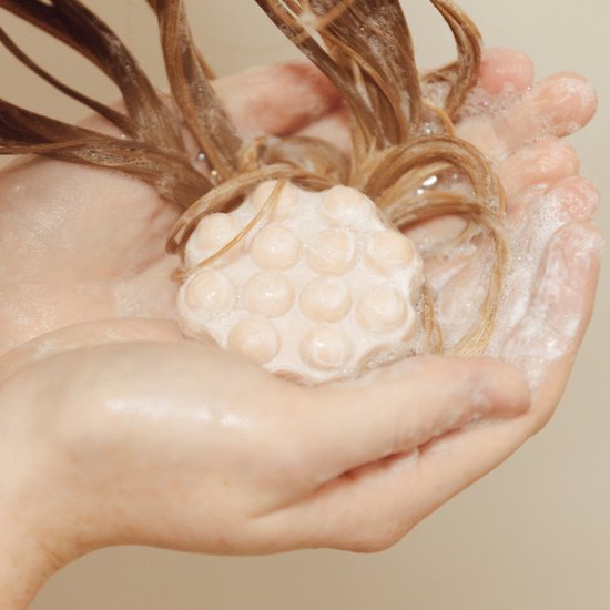 Shampoing solide Wondr rose pâle à picots dans des mains savonneuses avec des cheveux mouillés.