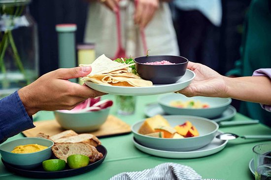 Een hand reikt een bord met crêpes en sla aan over een tafel vol eten.