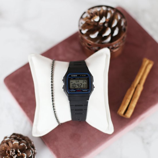 Montre numérique Casio F-91W noire sur coussin avec bracelet, pomme de pin et bâtons de cannelle.