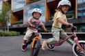 Twee lachende kinderen rijden op een loopfiets. Jongen op oranje-zwarte loopfiets, meisje op roze loopfiets.