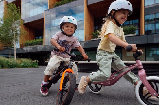 Twee lachende kinderen rijden op een loopfiets. Jongen op oranje-zwarte loopfiets, meisje op roze loopfiets.