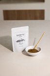 Witte zak met 'SHINYU hojicha' en een afbeelding van Mt. Fuji. Naast de zak een wit schaaltje met hojicha poeder en een bamboe schepje.